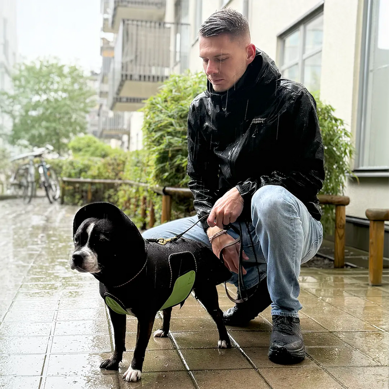 A man in a black rain jacket crouches next to a black medium sized dog wearing a black dog raincoat with a green belly wrap, on a wet tiled outdoor backyard. The rain is pouring down.