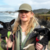 Blond woman in cap holding two small black mixed terrier dogs in her arms. Both dogs are wearing black dog raincoats, each with a green and a red belly wrap. The background is scenic by the ocean and high cliffs.