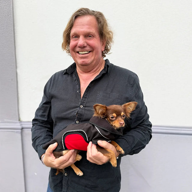 Man holding a chihuahua wearing a black modular raincoat with a removable red belly wrap against a white house wall background