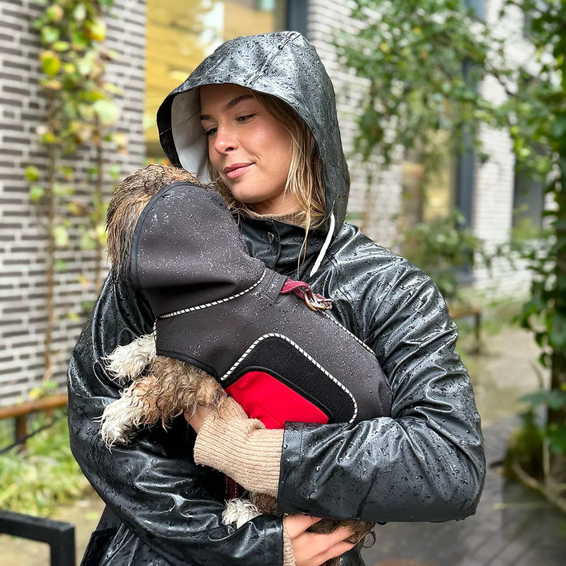 A blonde woman in a black raincoat holds a small dog wearing a black raincoat with a red belly wrap in the rain outdoors in front of a building.