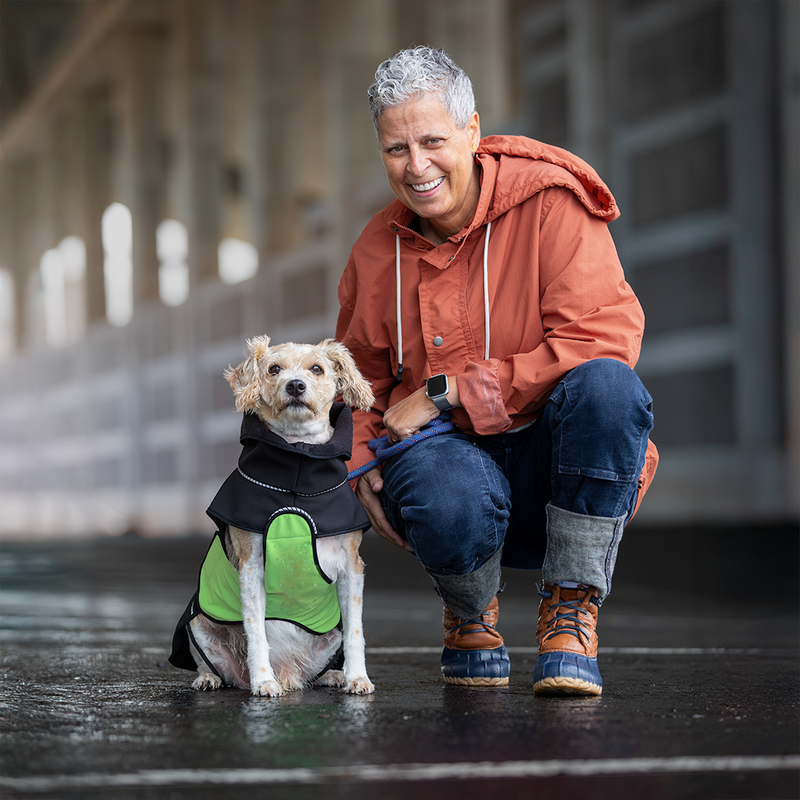 A smiling woman crouches beside a white medium sized dog on a wet pavement in an outdoor covered walkway. The woman wears an orange hooded jacket, blue jeans, and orange-and-blue boots, and the dog wears a black raincoat with a green belly wrap.