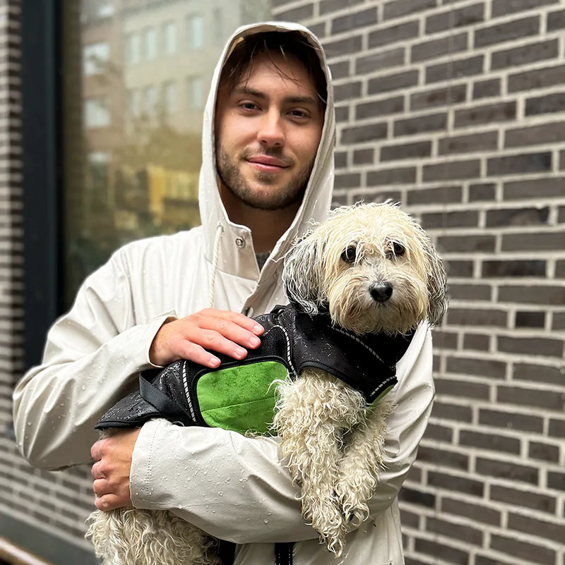 A young man in a beige hooded rain jacket holds a small fluffy dog wearing a black raincoat with a green belly wrap, standing against a gray brick wall. They are soaked by the rain.